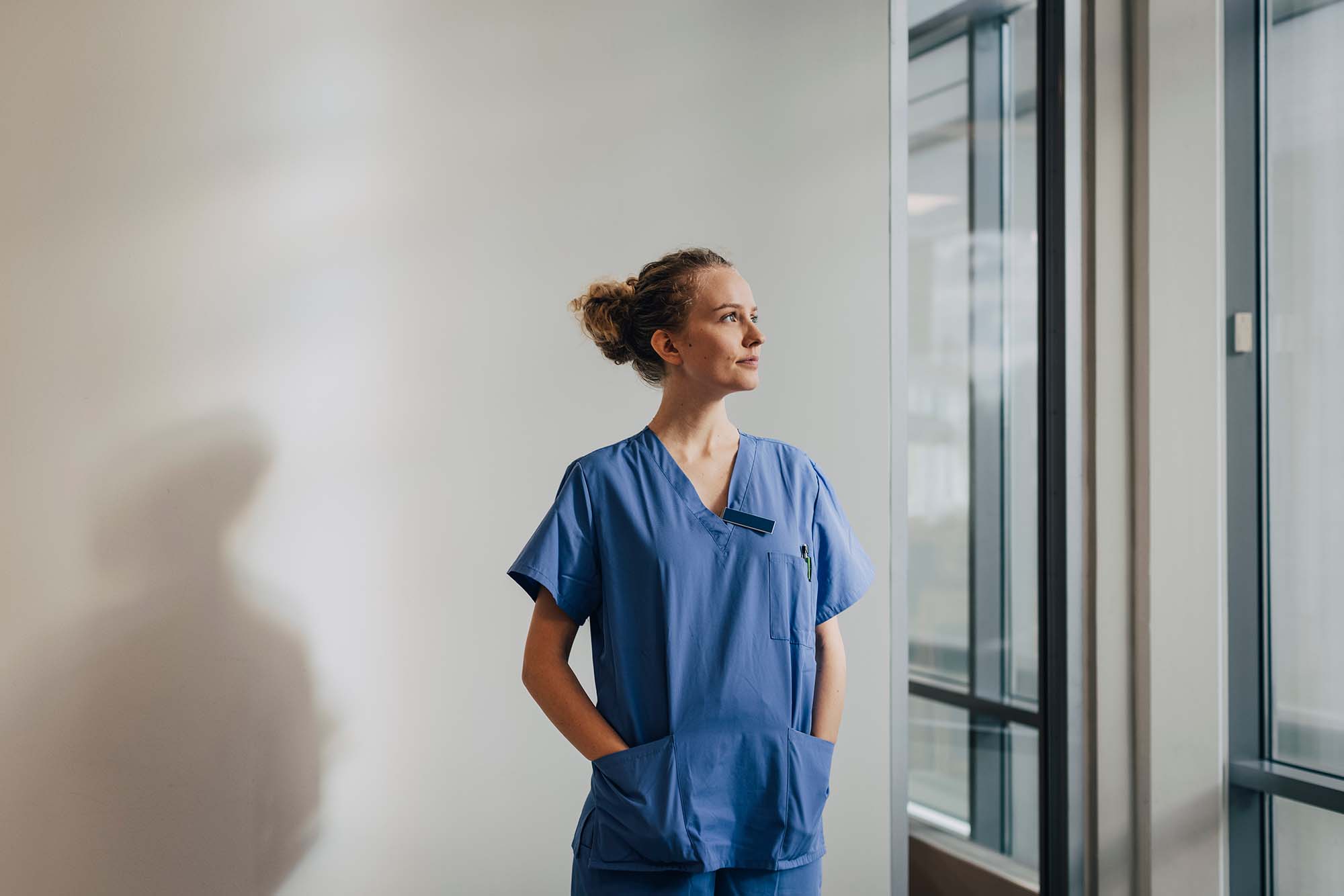 En kvinnlig sjuksköterska med blåa arbetskläder tittar ut genom fönstret