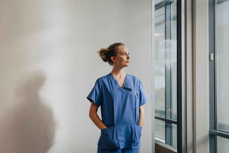 A female nurse wearing blue scrubs looking out through a window.